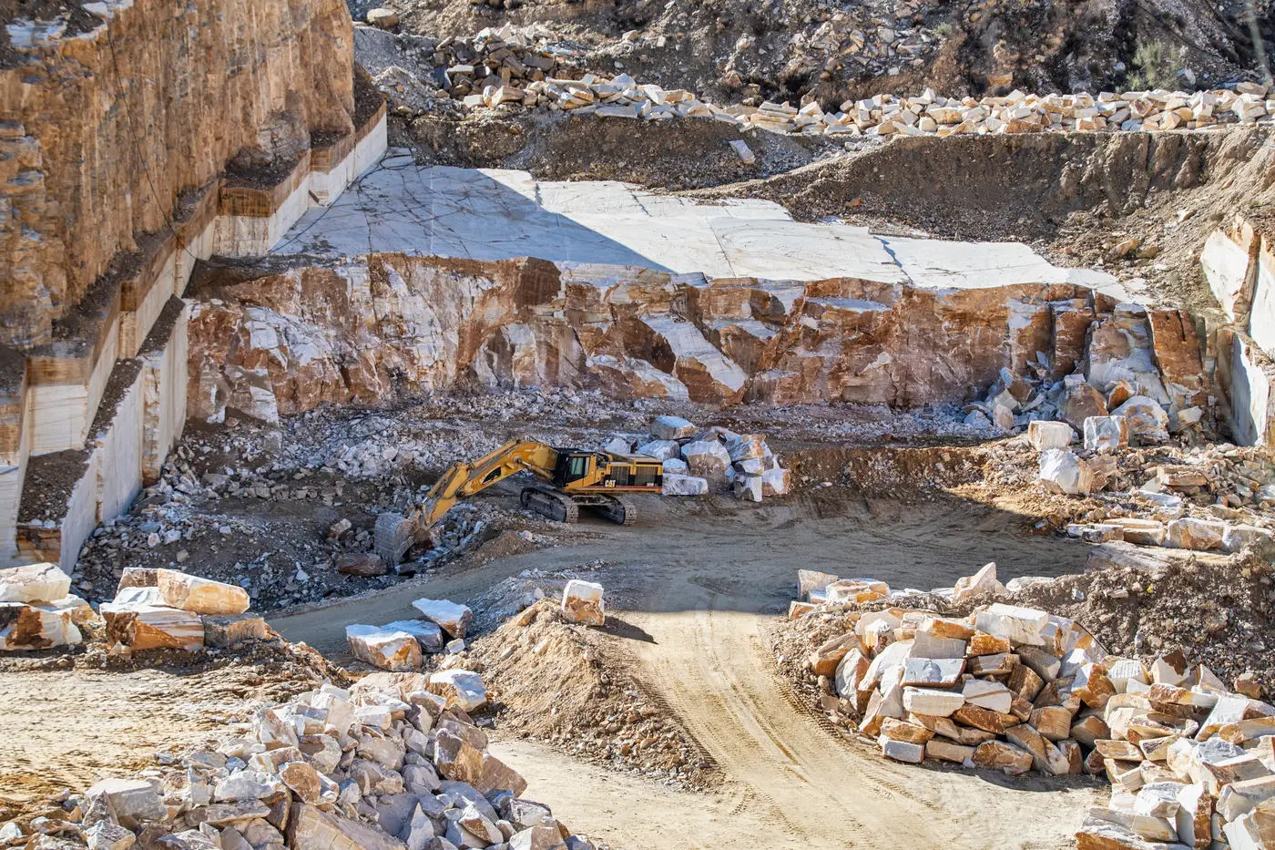 A yellow excavator works in a light-colored stone quarry surrounded by rubble.